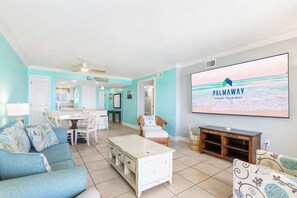 A living room with a TV displaying 'PALMAWAY VACAY YOUR WAY,' a dining area with a table and chairs, and a kitchen in the background. - The Big Surf II - Gulf View - Beach, Pool, and Fun (Gulf Shores)