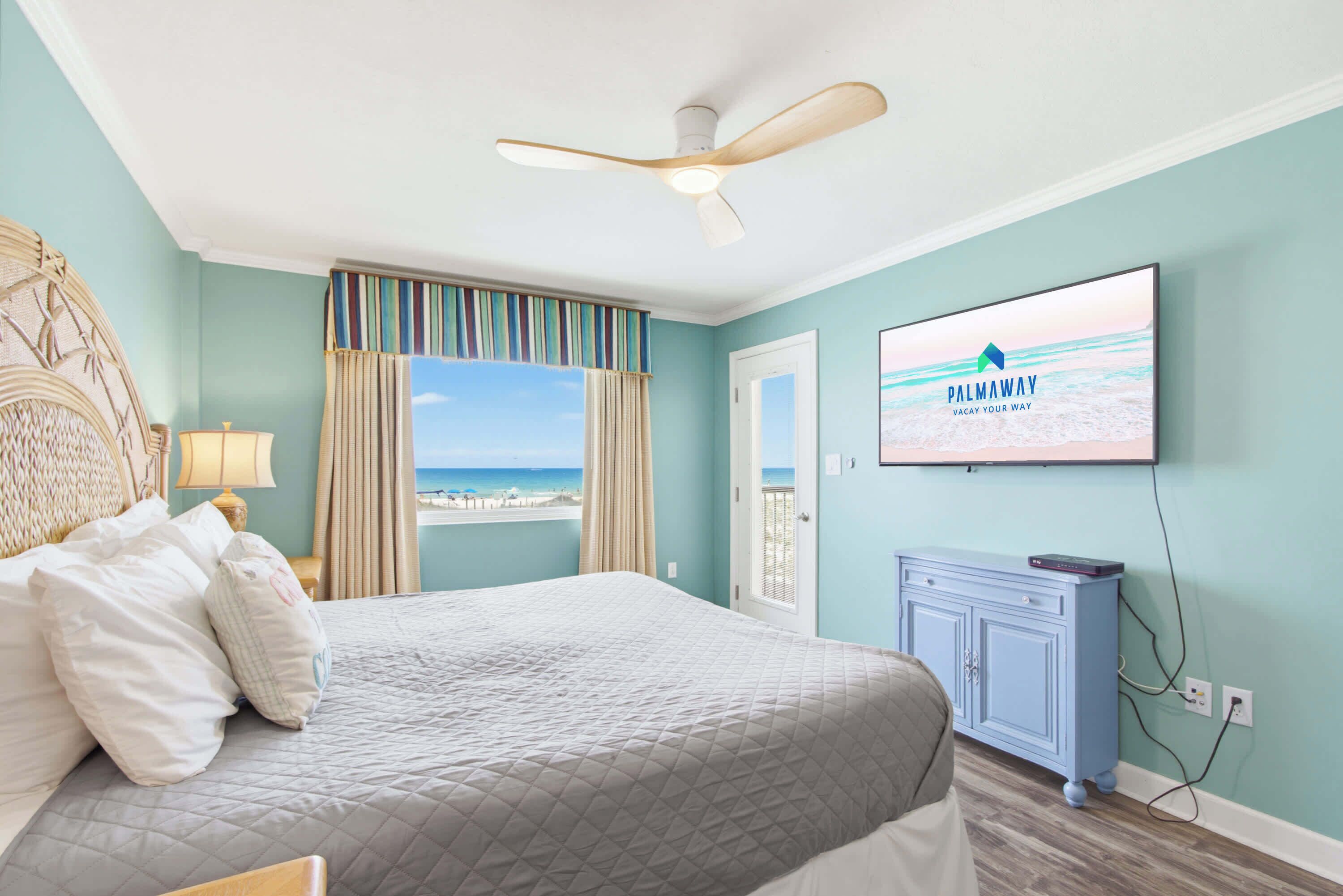 A bedroom with a bed, a ceiling fan, a television, and a view of the beach.