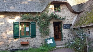 A rustic stone building with a thatched roof, a wooden door, and a small window with green shutters. - Verte Printanière Guest Room – Upstairs with Shared Terrace & Garden, Wi-Fi (Saint-Lyphard)