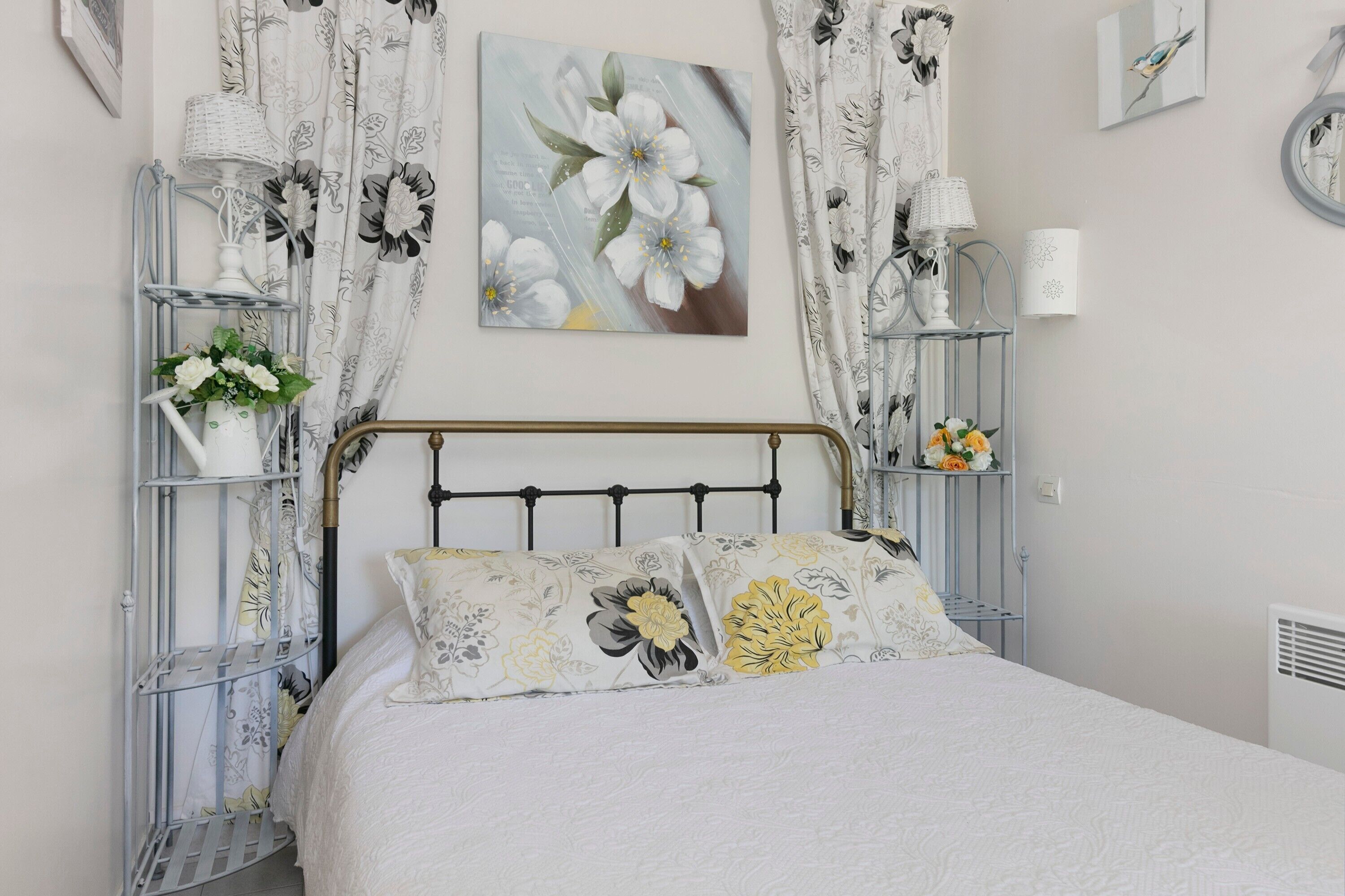 Een slaapkamer met een bed, bloemen gordijnen, een wandlamp en een plank met een bloevas.