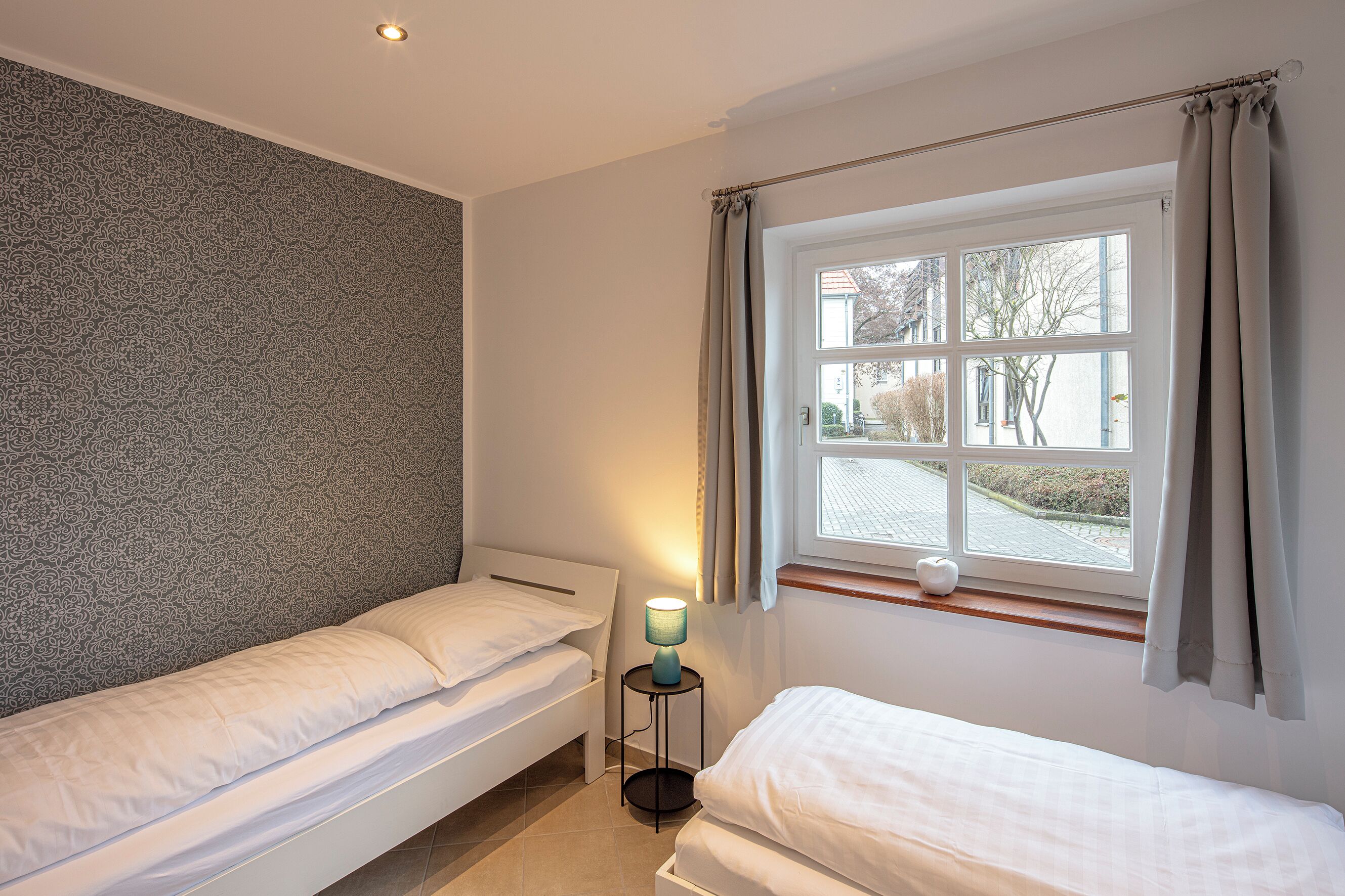 Ein Schlafzimmer mit zwei Einzelbetten, einem Nachttisch mit Lampe, einem Fenster mit Vorhängen und einer Wand mit Muster.