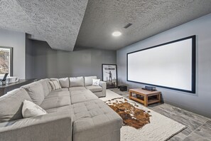 Ein modernes Wohnzimmer mit einer Eckcouch, einem großen Flachbildfernseher, einem Kuhfellteppich und einem Projektor auf einem Holztisch.