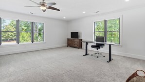 Une pièce spacieuse dotée d’un ventilateur de plafond, d’un bureau avec une chaise, d’un téléviseur posé sur un meuble en bois et de grandes fenêtres équipées de stores.