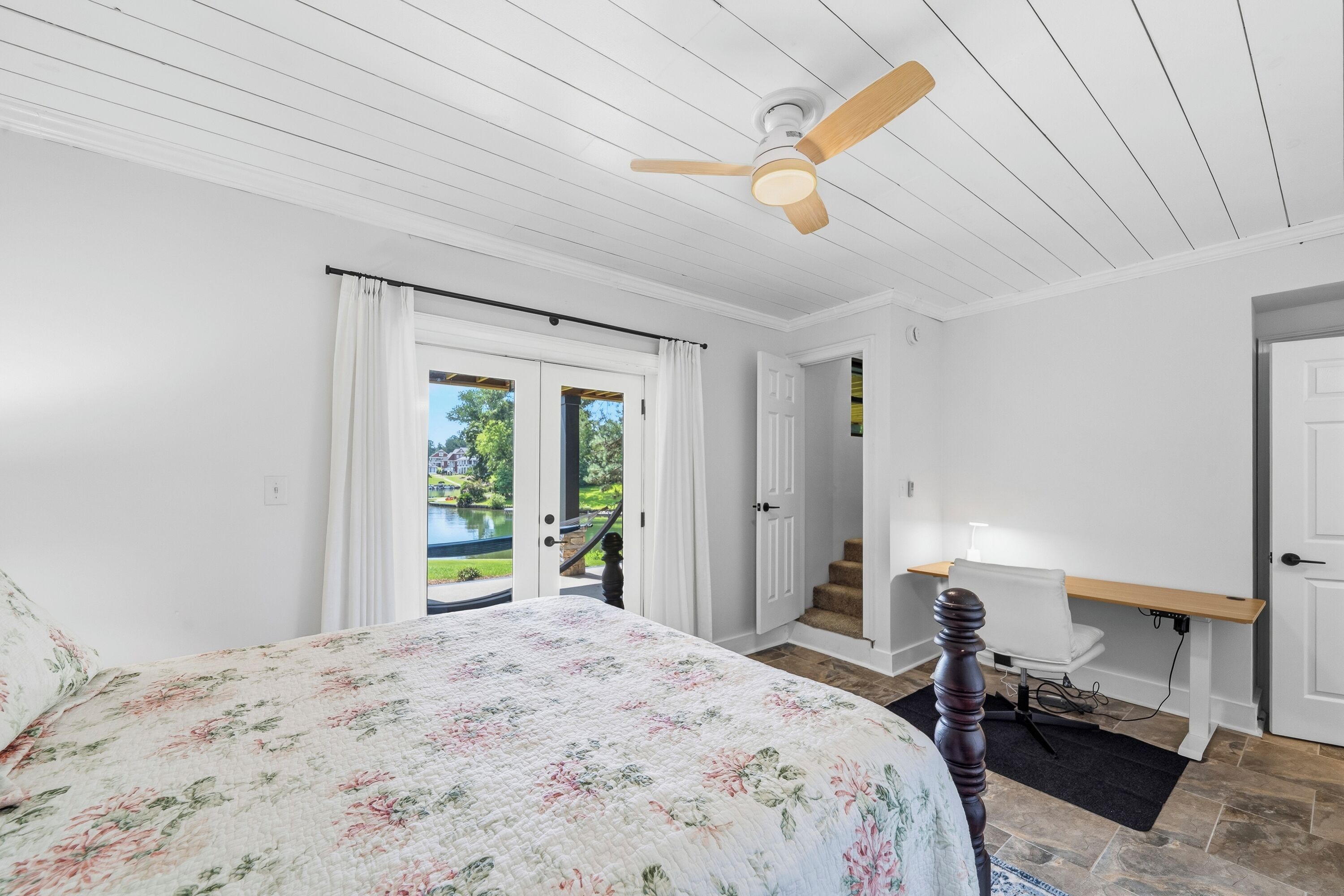 Un dormitorio con cama, ventilador de techo, escritorio y vistas a un área exterior a través de una puerta.