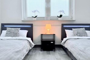 A hotel room with two beds, a bedside table with a lamp, and a window with blinds.