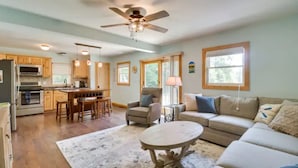 A spacious living area with a sofa, coffee table, and a dining table with chairs. There is a kitchen area with wooden cabinets and a ceiling fan. - Lakefront 3-bedroom chalet with private dock (Louisa)