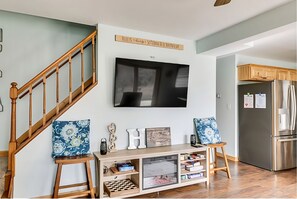 Un salon avec un escalier en bois, un téléviseur à écran plat, une console ornée d’objets décoratifs et un coin cuisine avec un réfrigérateur.