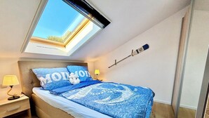 Ein Schlafzimmer mit Oberlicht, einem Bett mit blauer Bettwäsche, einer Nachttischlampe und einem an der Wand befestigten Besen.