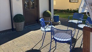 Ein Gebäude mit einer braunen Tür, einem kleinen Fenster und einer überdachten Terrasse mit blauen Stühlen und einem Korbwäscher.