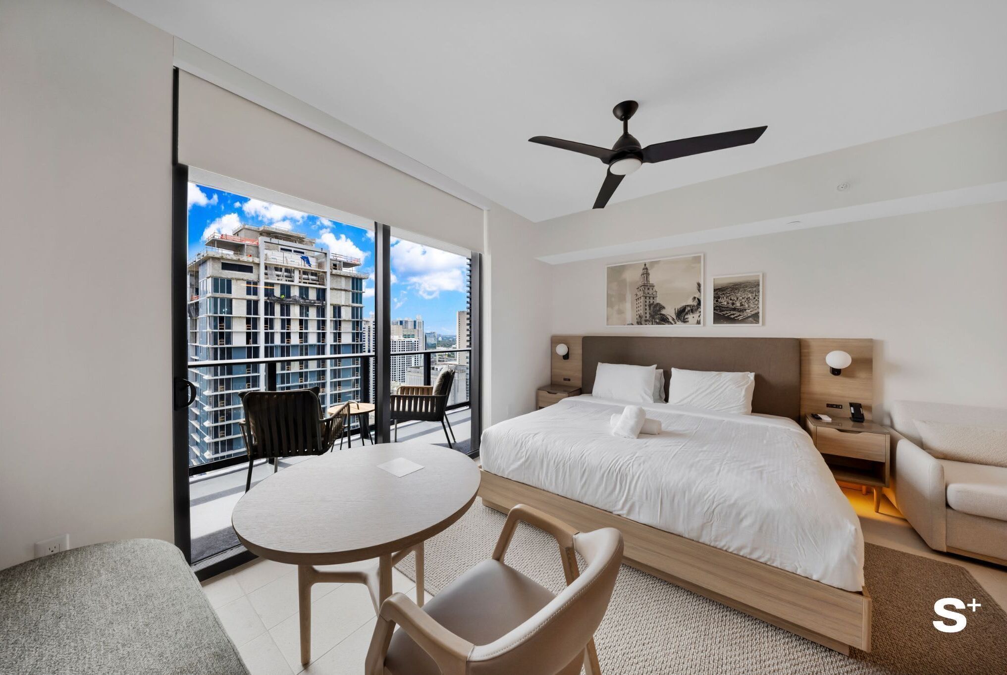 Una habitación de hotel moderna con una cama grande, un ventilador de techo, un balcón con mesa y sillas, y vistas a la ciudad.