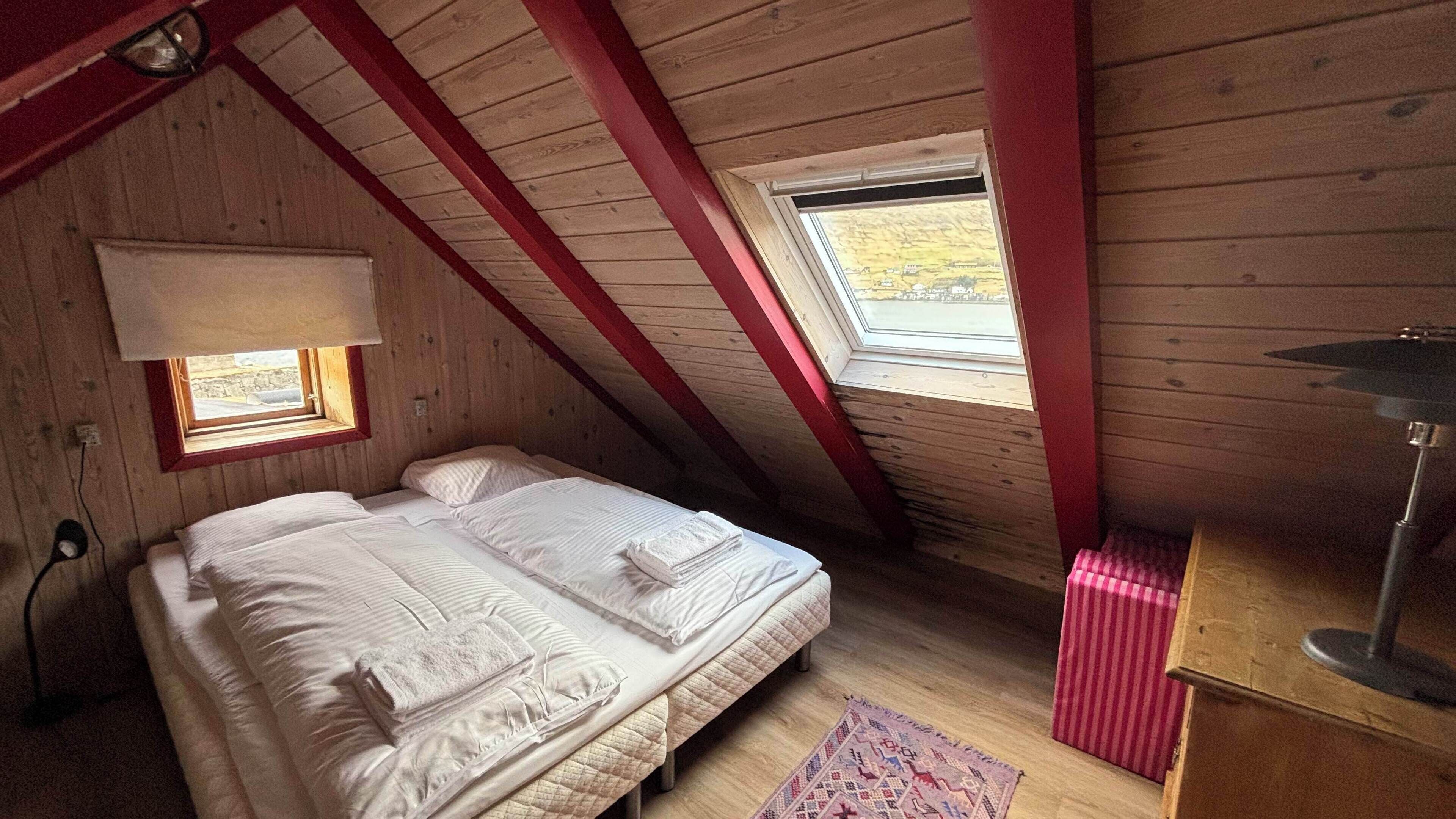 Une chambre avec un plafond en pente, un lit recouvert de draps blancs, une petite fenêtre et une lampe.