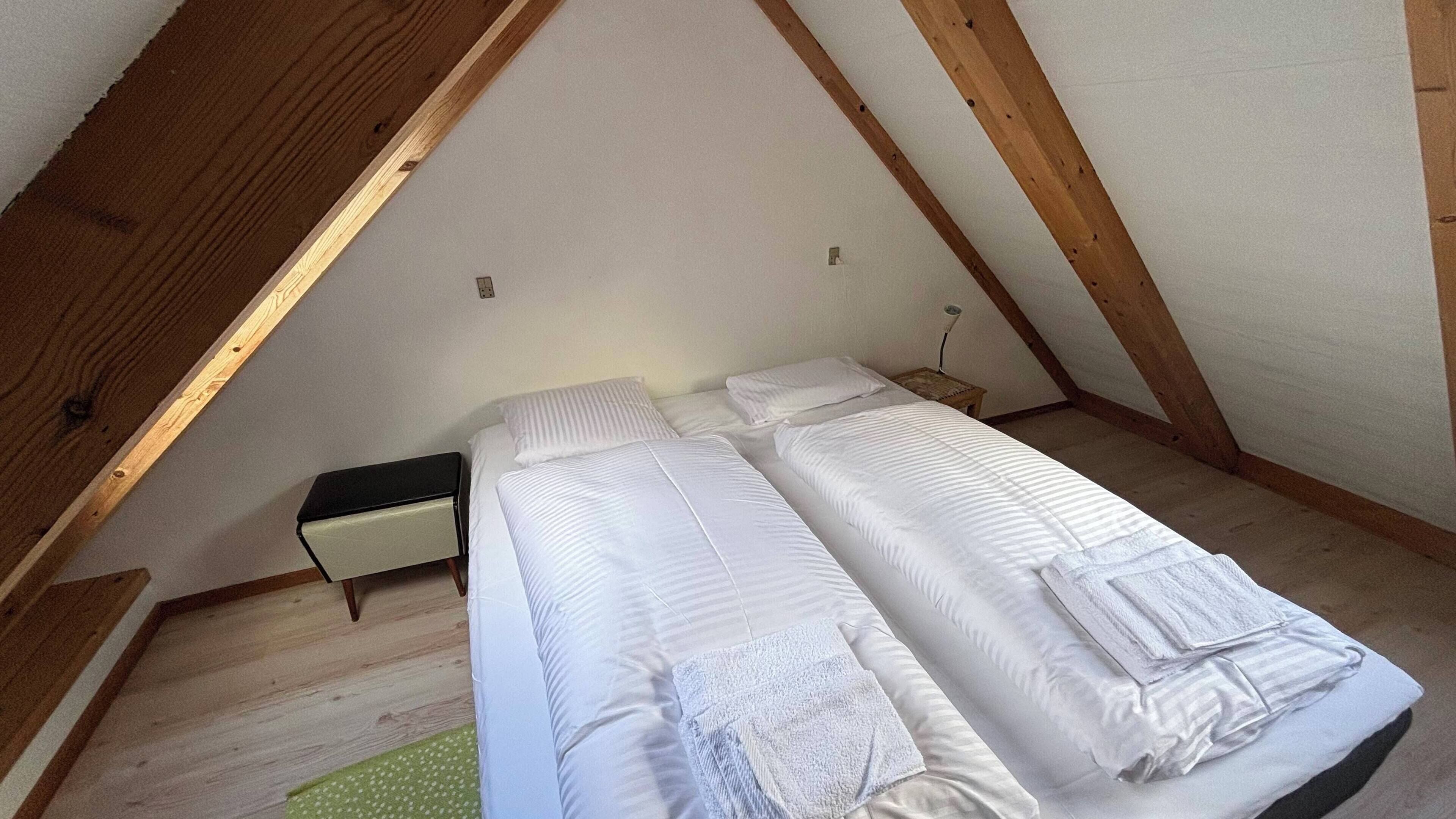 Un petit logement composé d’une seule pièce, doté d’un plafond en pente et de poutres en bois, comprenant deux lits avec du linge de lit blanc et une table de chevet.