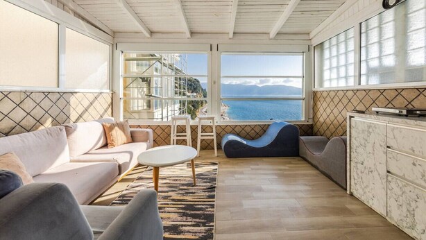 A modern living room with a sofa, a small table, and a bean bag chair. Large windows offer a view of the sea and buildings. - Vista Mare Horizon | Patio with Amazing Seaview by PikHost (Vlorë)