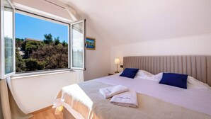 Hotellihuone, jossa on sänky, yöpöytä, lamppu, ikkuna, josta avautuu näkymä puihin ja rakennukseen, sekä taulu seinällä.