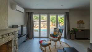A living room with a stone fireplace, a flat-screen TV, a comfortable armchair, and a view of a swimming pool. - Villa Omega - Two Bedroom Villa, Sleeps 4 (Ditiki Mani)