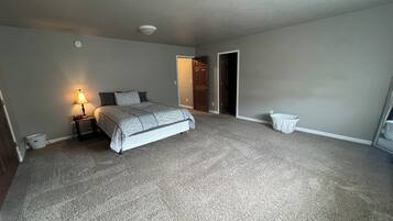 A bedroom with a bed, a bedside lamp, a carpeted floor, a door, and a small trash bin.