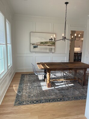 A dining area with a wooden table, clear chairs, a patterned rug, and a painting of a coastal scene. - Masters Getaway in Evans, GA (Evans)