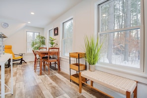 A room with a dining table, chairs, a bench, potted plants, and large windows with a view of trees.