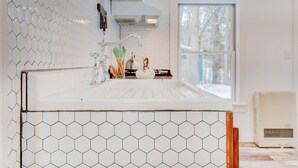 A kitchen with a white countertop, hexagonal tile backsplash, and a window with a view of trees. - Tiny Acres: A Storybook Tiny Home, 4 Mi. to CDP (Milford)