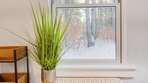 A window with a view of a snowy forest, a wooden chair, and a potted plant on a woven mat. - Tiny Acres: A Storybook Tiny Home, 4 Mi. to CDP (Milford)