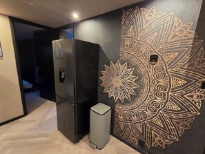 A modern kitchen with a black refrigerator, a grey trash bin, and a decorative wall with a mandala pattern.