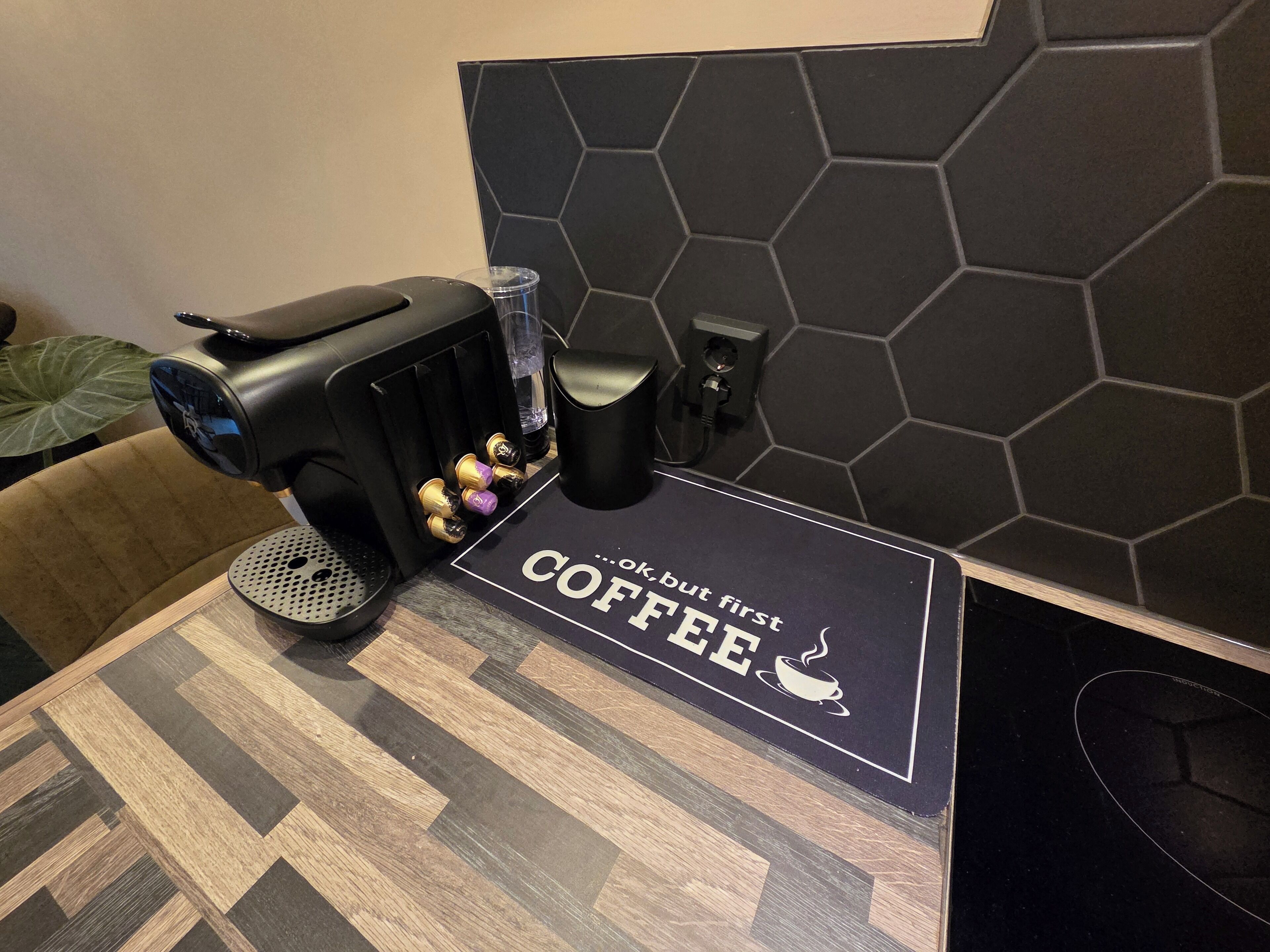 A coffee machine, a cup, and a coaster with 'Coffee, but first' text on a table.