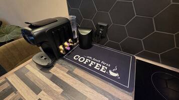 A coffee machine, a cup, and a coaster with 'Coffee, but first' text on a table.