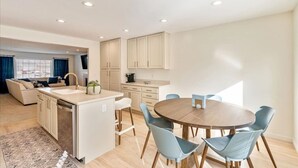 Cozinha moderna com ilha central, sala de jantar com mesa e cadeiras redondas, e sala de estar com sofá e máquina de café.