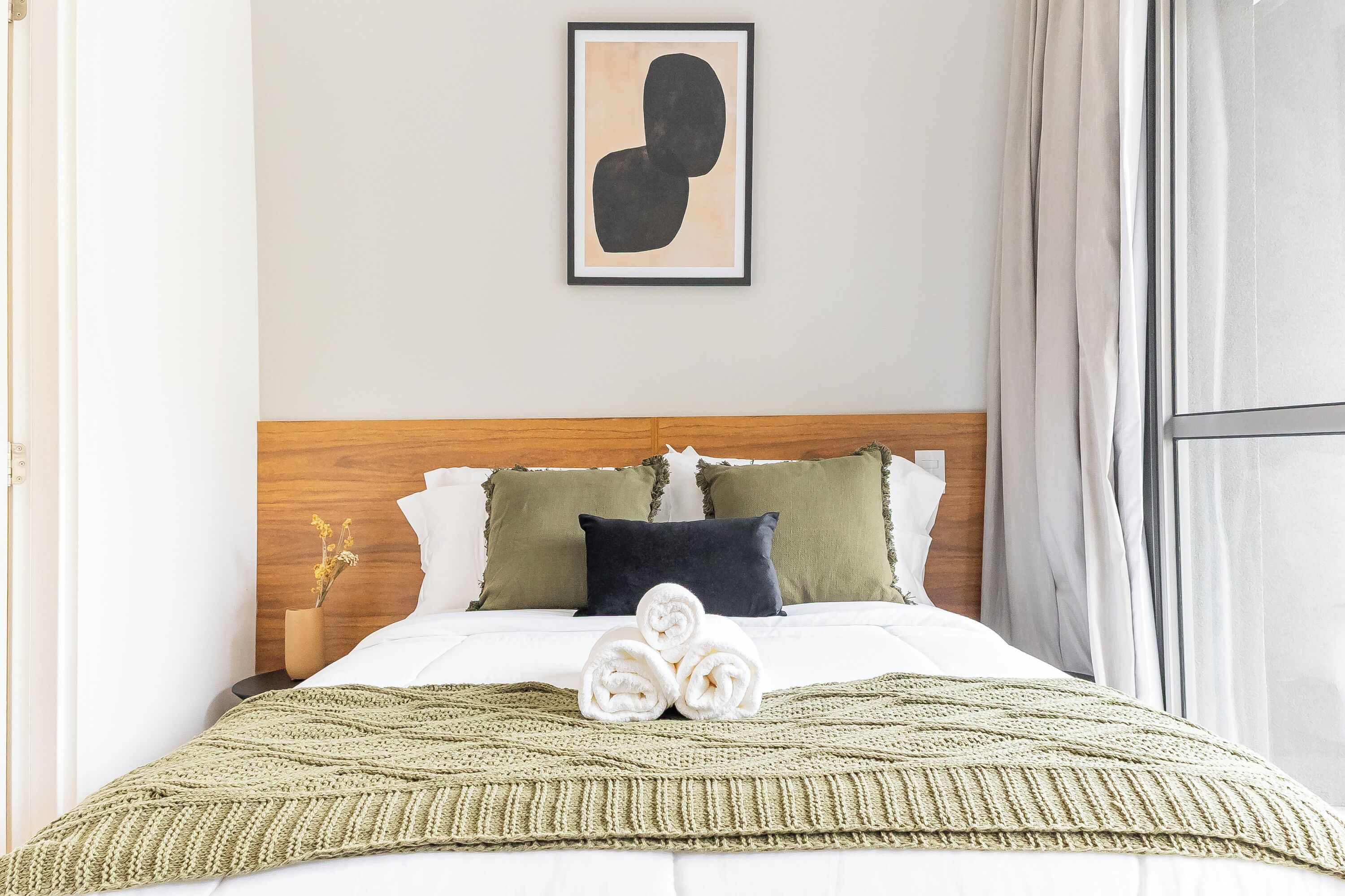 Ein ordentlich bezogenes Bett mit grüner Decke, einem gerahmten abstrakten Kunstwerk und einem hölzernen Kopfteil.