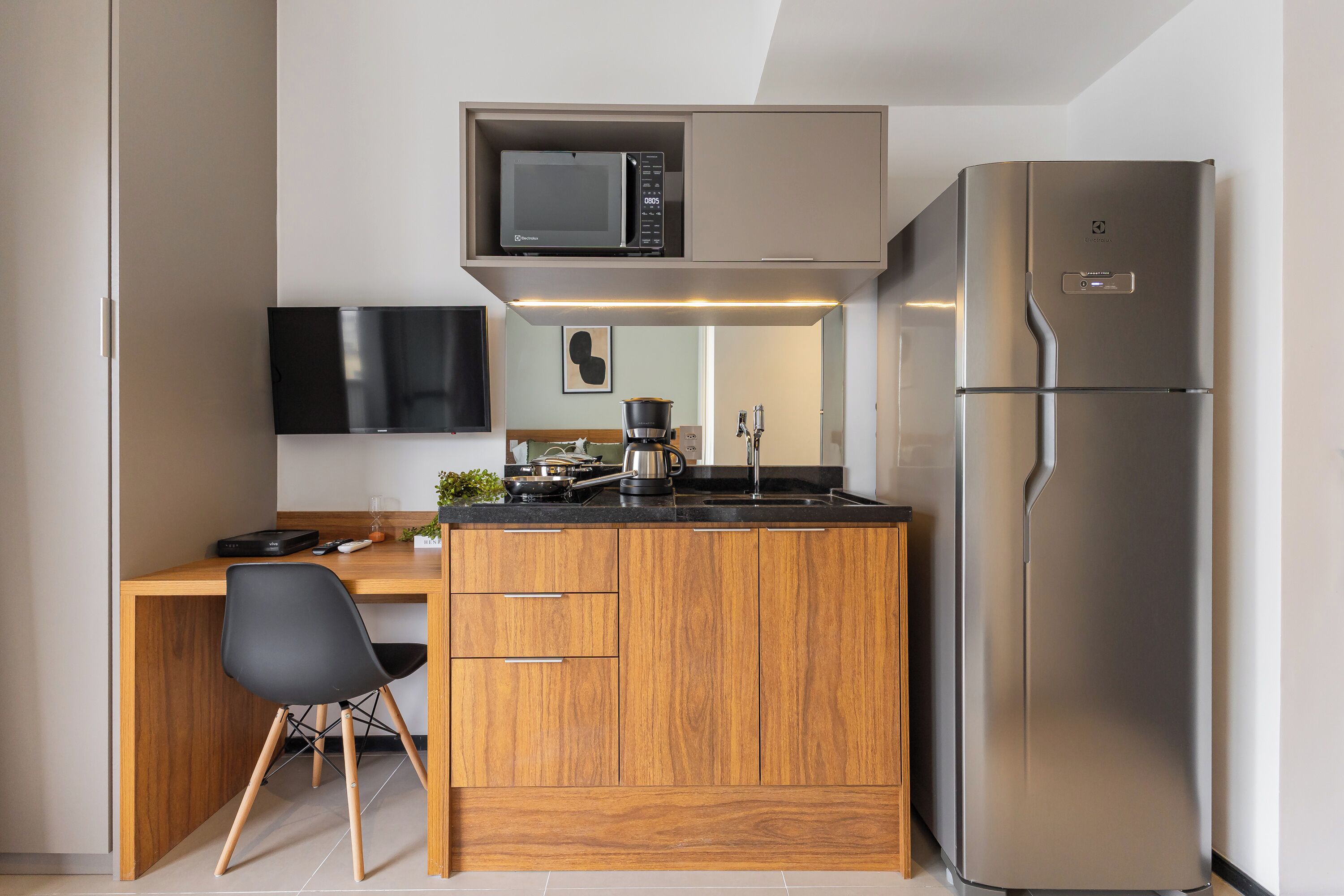 Una cucina compatta con forno a microonde, frigorifero, pensili in legno, una sedia nera e una TV a schermo piatto fissata al muro.