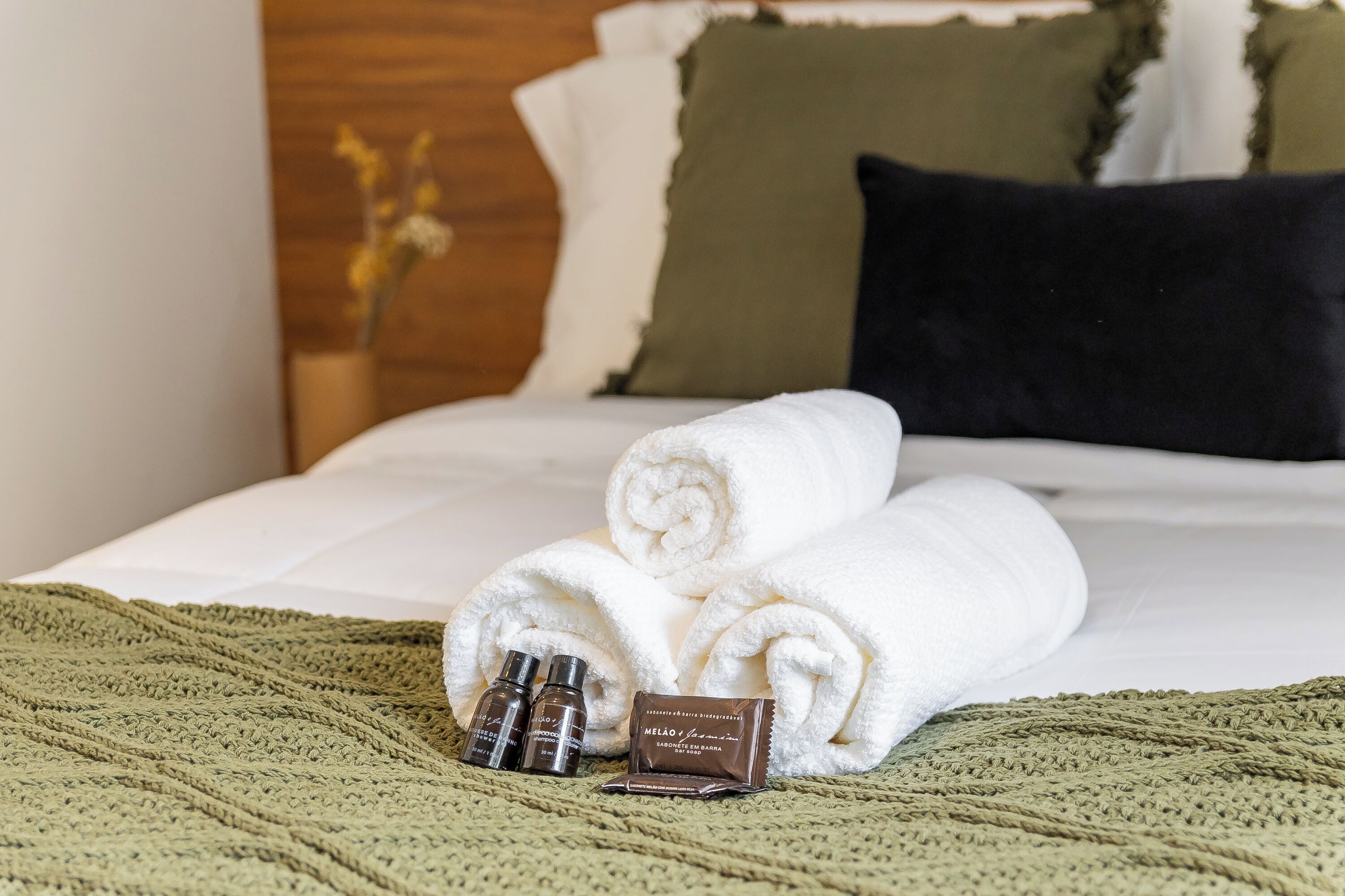 A neatly made bed with rolled white towels, a green knitted blanket, and two small bottles of lotion on the bed.