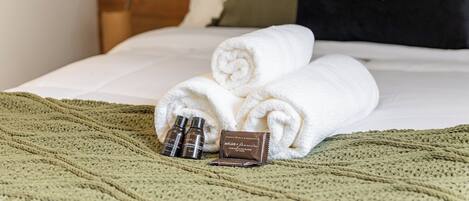 A neatly made bed with rolled white towels, a green knitted blanket, and two small bottles of lotion on the bed.