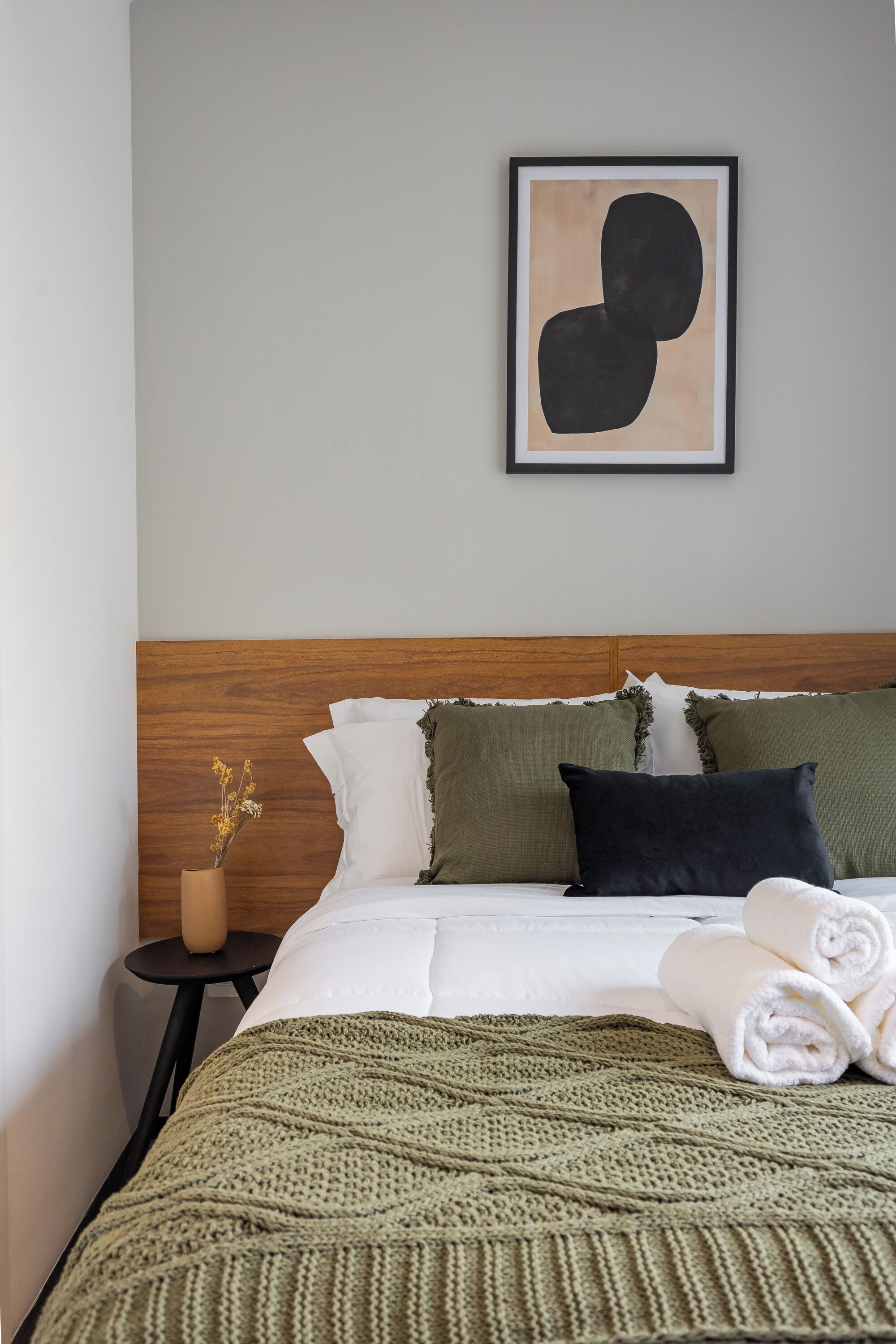 A neatly made bed with a green knitted blanket, a wooden headboard, and a framed abstract artwork on the wall.