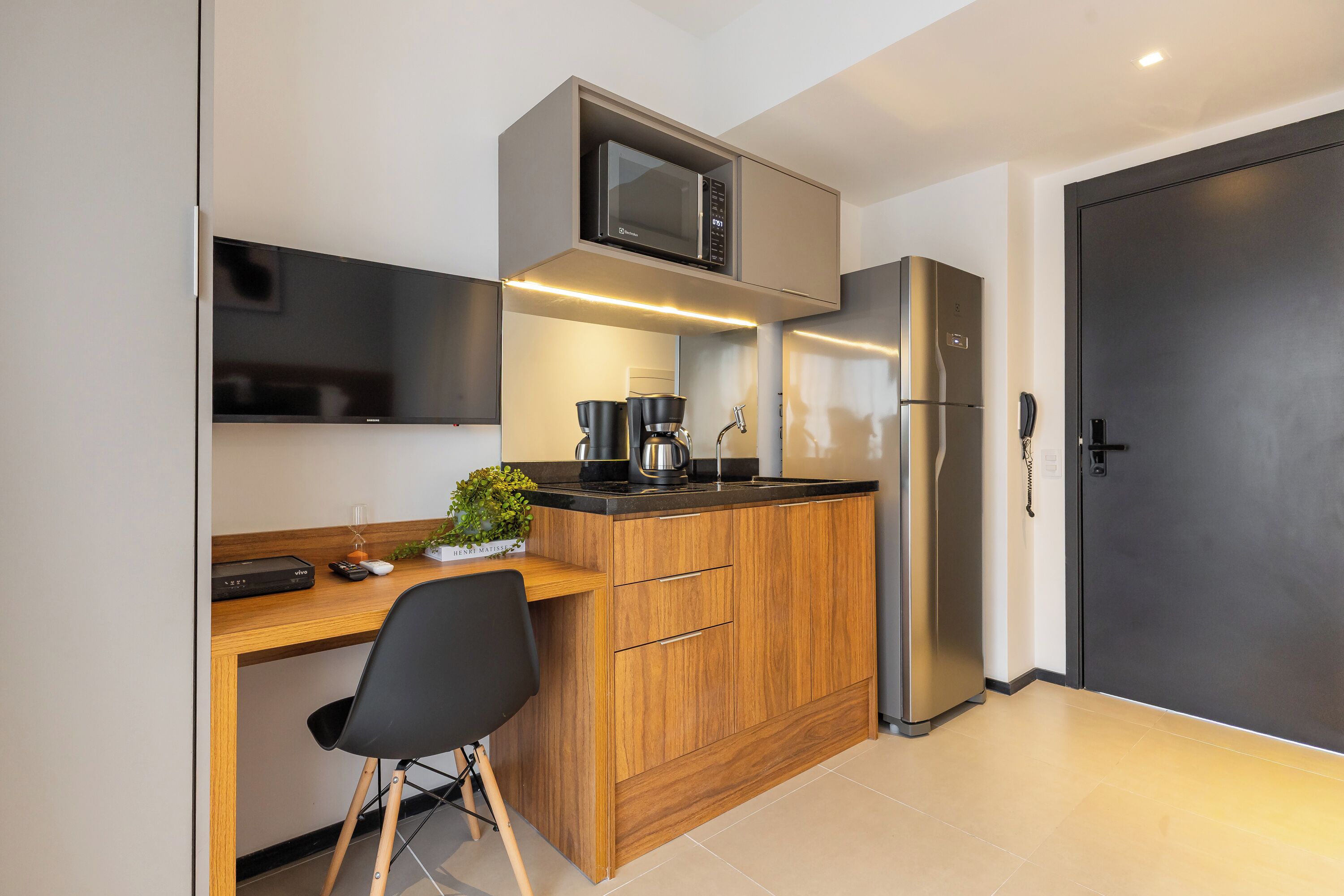 A compact kitchen with a microwave, coffee maker, and refrigerator, a wooden desk with a chair, and a dark door.