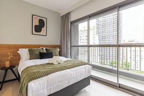A modern hotel room with a large bed, a bedside table with a vase, a framed picture on the wall, and a view of high-rise buildings. - StayHome Consolação Gym and Balcony (São Paulo)