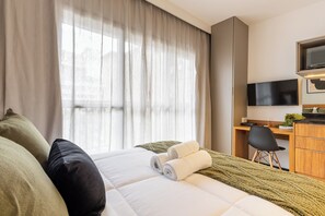 A hotel room with a bed, a desk, a chair, a television, and a window with curtains. - StayHome Consolação Gym and Balcony (São Paulo)