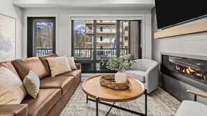 A modern living room with a leather sofa, a round coffee table, a fireplace, and a large window with a view of a building and mountains. - 3 Bed/3 Bath Cozy Mountain Retreat (Canmore)