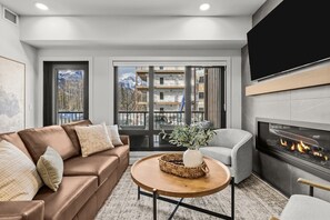 Ein modernes Wohnzimmer mit Ledersofa, rundem Couchtisch, Kamin und großem Fenster mit Blick auf ein Gebäude und Berge.