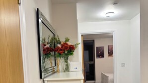 Un corridoio con un armadietto bianco, uno specchio e un vaso con fiori. Si vede una porta a sinistra e una stanza sullo sfondo.
