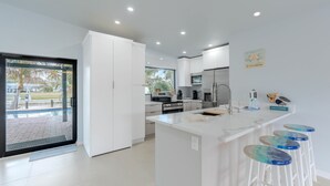 Cocina moderna con armarios blancos, una isla central con taburetes y un refrigerador de acero inoxidable, con vista a una piscina exterior.
