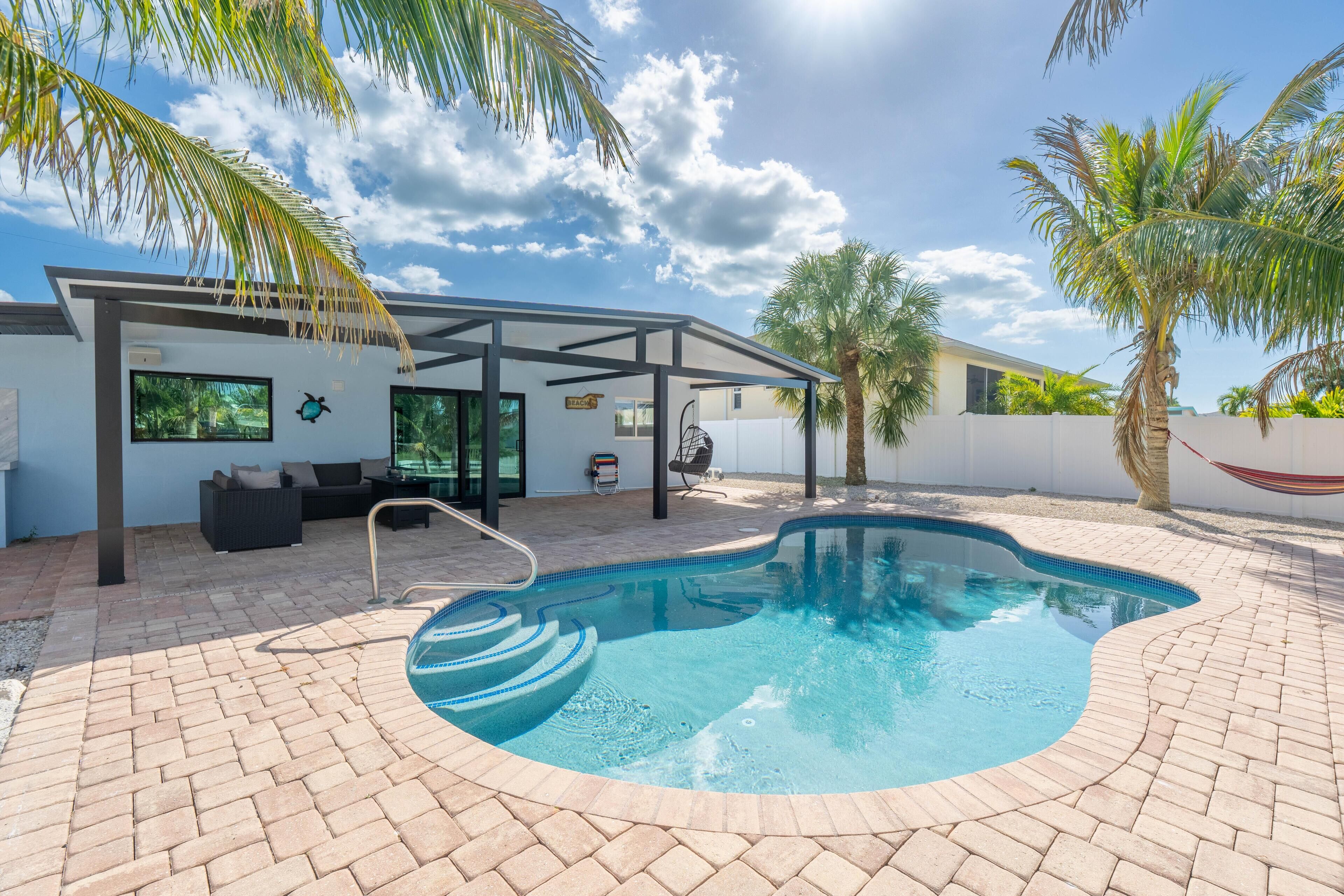 A swimming pool with a sunshade, a lounge area with a sofa, and palm trees.