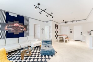 Un salon moderne avec un sol à carreaux, des canapés blancs, un pouf bleu et un coin repas comprenant une table et des chaises.
