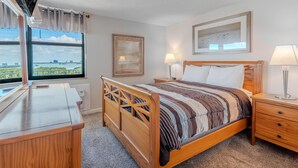 Une chambre d’hôtel avec un lit en bois, deux tables de chevet, un bureau, une télévision et une grande fenêtre donnant sur la ville.