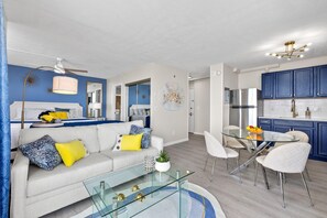 A modern living room with a glass coffee table, a sofa with yellow and blue cushions, a dining area with a glass table and white chairs, and a kitchen with blue cabinets and stainless steel appliances. - Estero Beach & Tennis 1102-a (Fort Myers Beach)