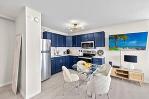 A modern kitchen with blue cabinets, stainless steel appliances, a glass dining table, and a beach-themed painting. - Estero Beach & Tennis 1102-a (Fort Myers Beach)