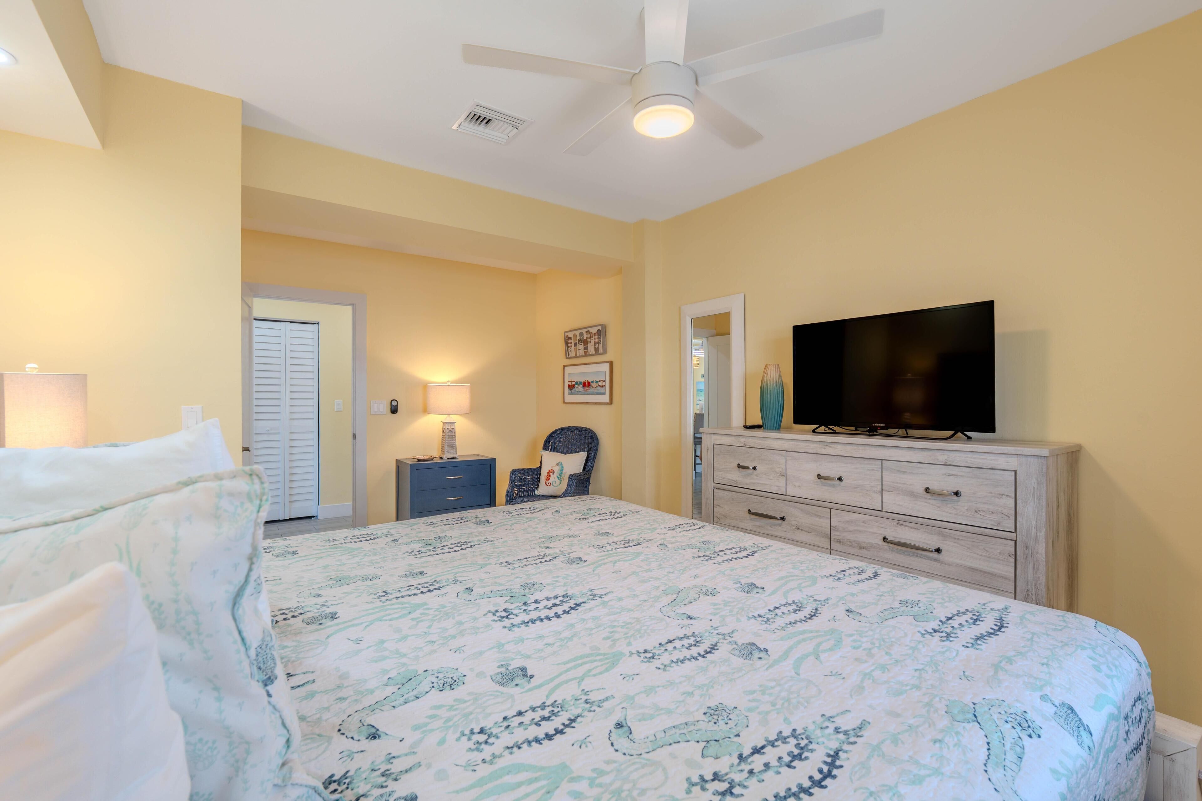 Un dormitorio con cama, televisión, cómoda, silla y puerta.