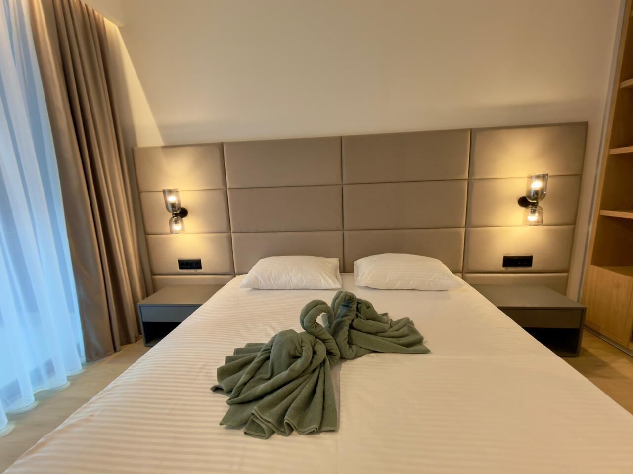 Una habitación de hotel con una cama grande, dos mesitas de noche, un cabecero y una mesita con lámpara.