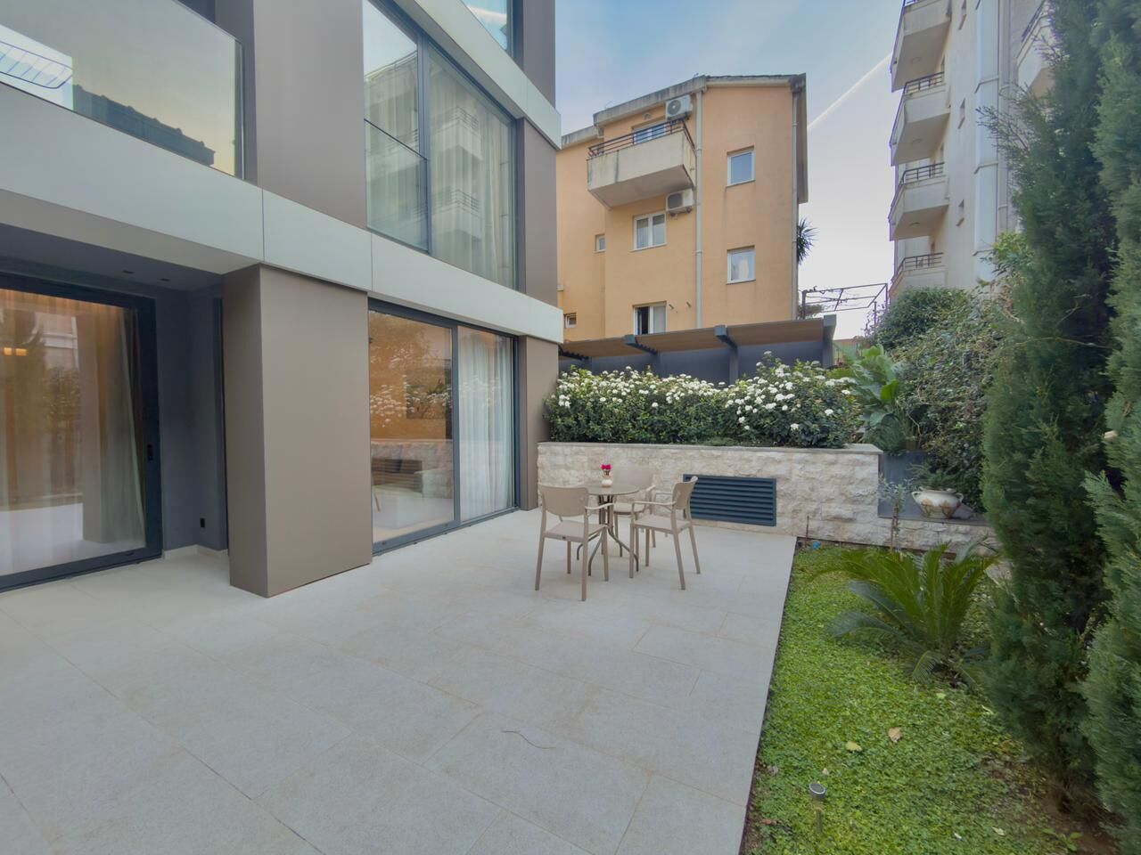 A modern building with a patio area featuring a table and chairs, surrounded by greenery and a stone wall.