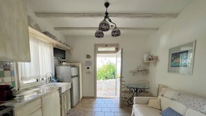 A compact kitchen with a refrigerator, stove, and sink. There is a dining area with a table and chairs, and a sofa with a striped pillow. A door leads to an outdoor patio. - Ostuni Green Hills App. 2 - Panoramic view, charm, veranda and shared jacuzzi (Puglia)