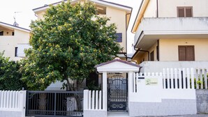 Ein begrenzter Eingangsbereich mit weißem Zaun, ein Baum mit gelben Blüten und ein Gebäude mit beiger Fassade und braunen Fensterläden.